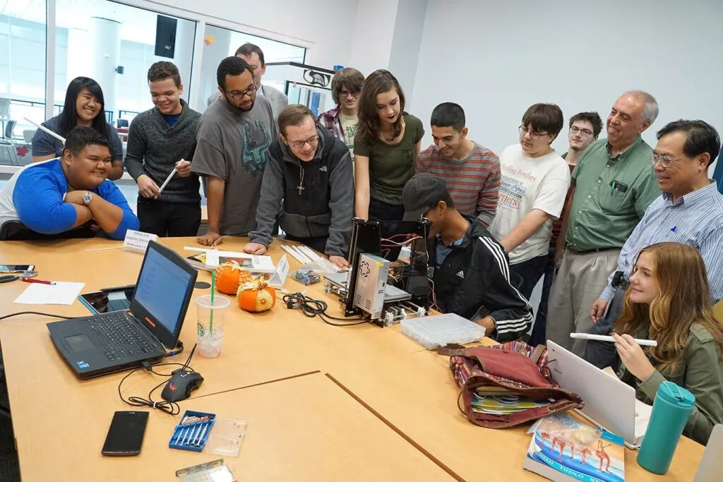 Group of students gathered around a table watching a student work on a project