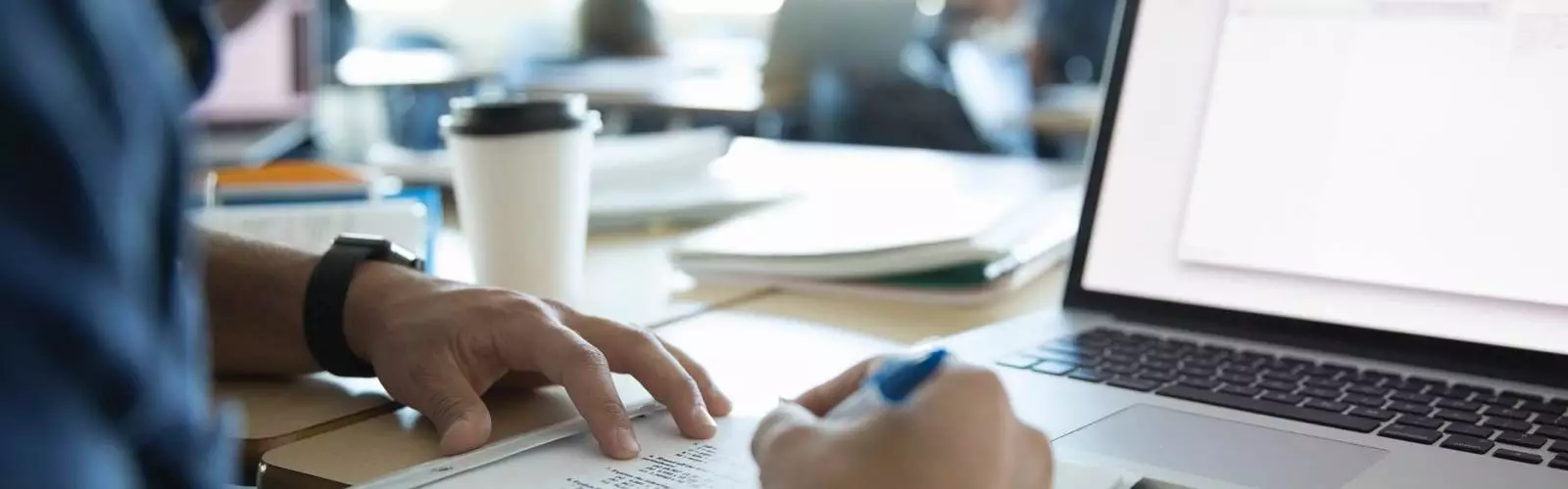 Person writing on paper while working on a laptop