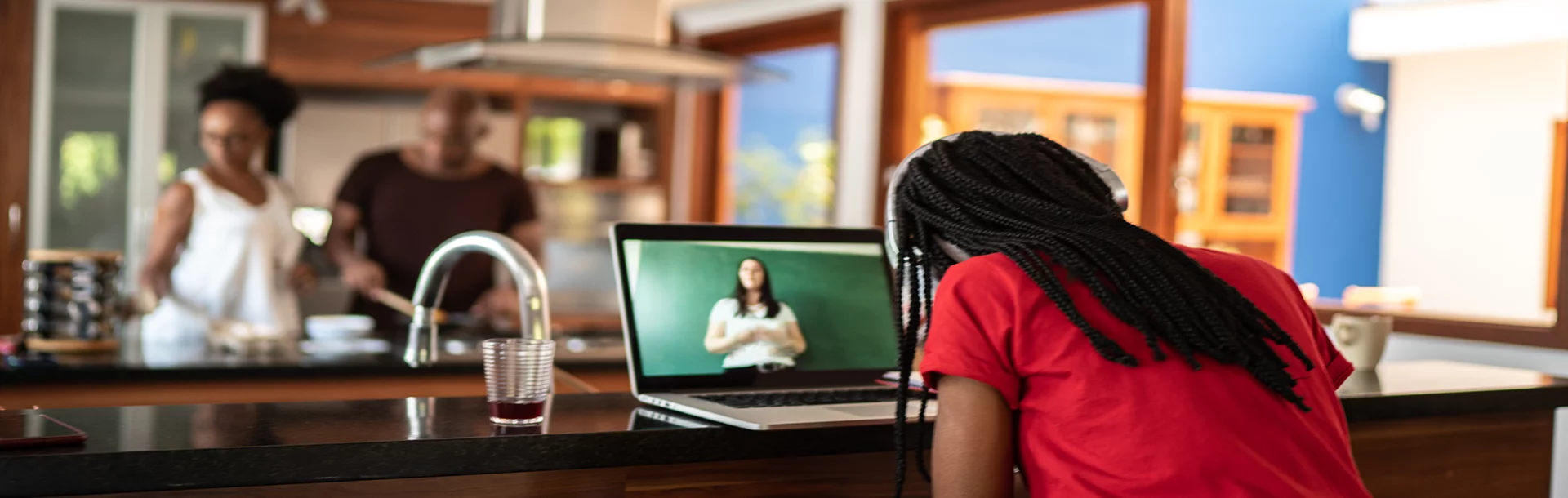 Woman sitting in the kitchen on her laptop taking an online class while someone cooks in the background