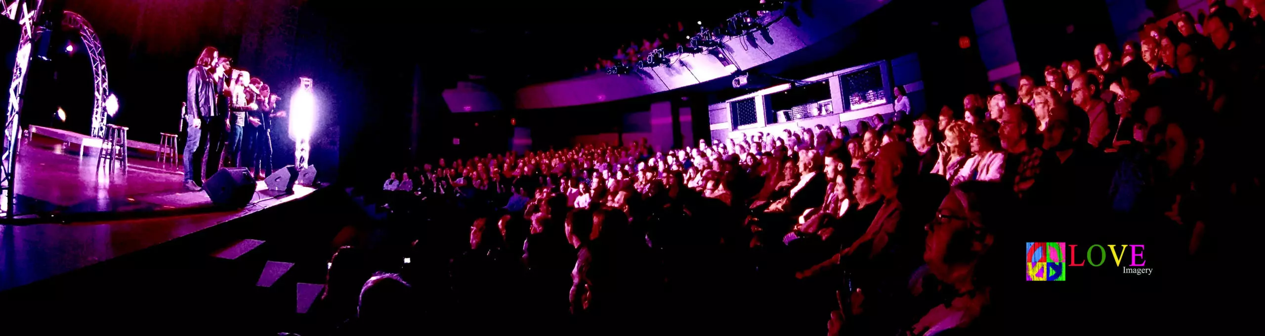 Interior of the Grunin Center theatre with a full audience, lighted stage and performers at the edge of the stage. Logo for Love Imagery includes peace sign.