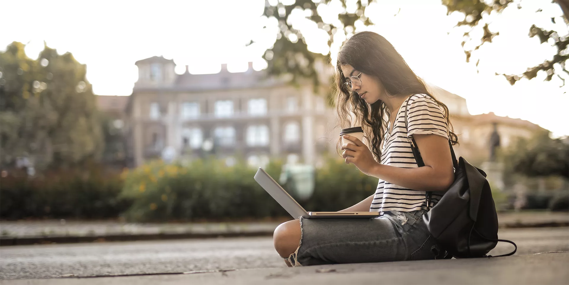 woman sitting on the ground on campus on her laptop while drinking coffee